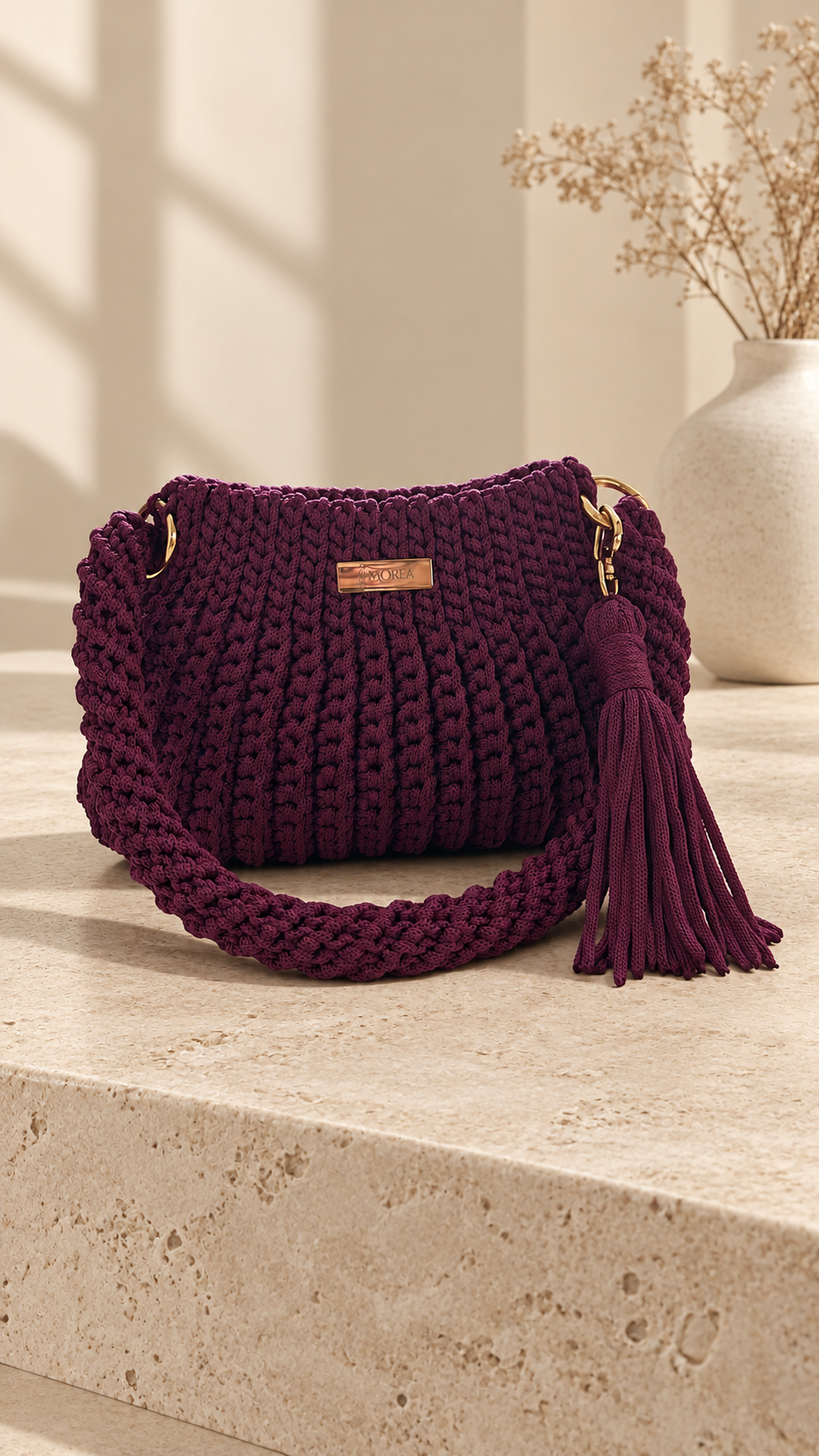 Burgundy crochet Jolie shoulder bag with gold hardware and a fringe tassel, on a stone ledge beside a vase of dried flowers.