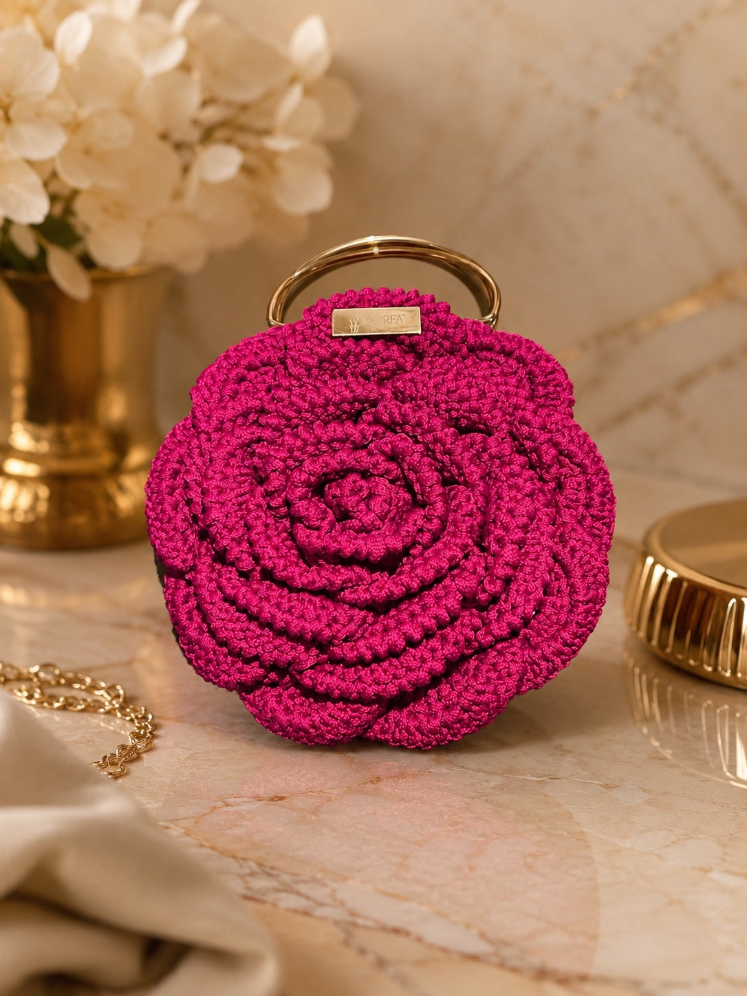 Pink crochet rose-shaped clutch bag with a gold handle and MOREA tag, styled on a marble surface with flowers and gold accessories.