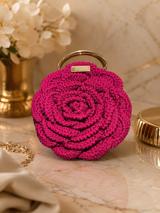 Pink crochet rose-shaped clutch bag with a gold handle and MOREA tag, styled on a marble surface with flowers and gold accessories.