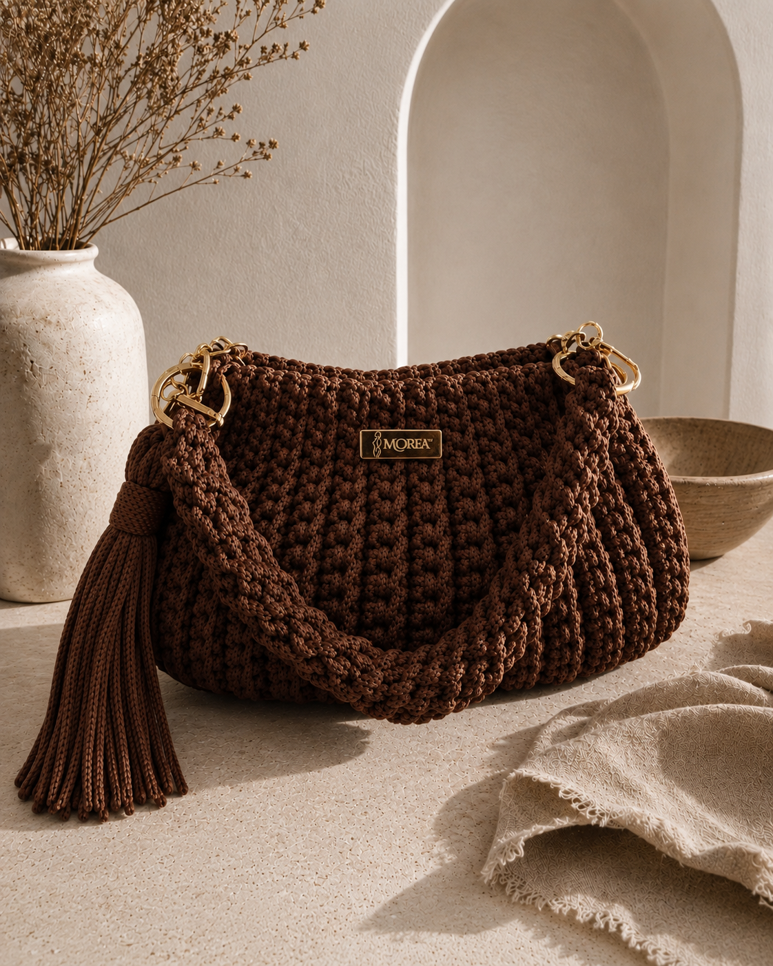 Brown crochet Jolie shoulder bag with a fringe tassel, styled beside a ceramic vase and linen cloth.