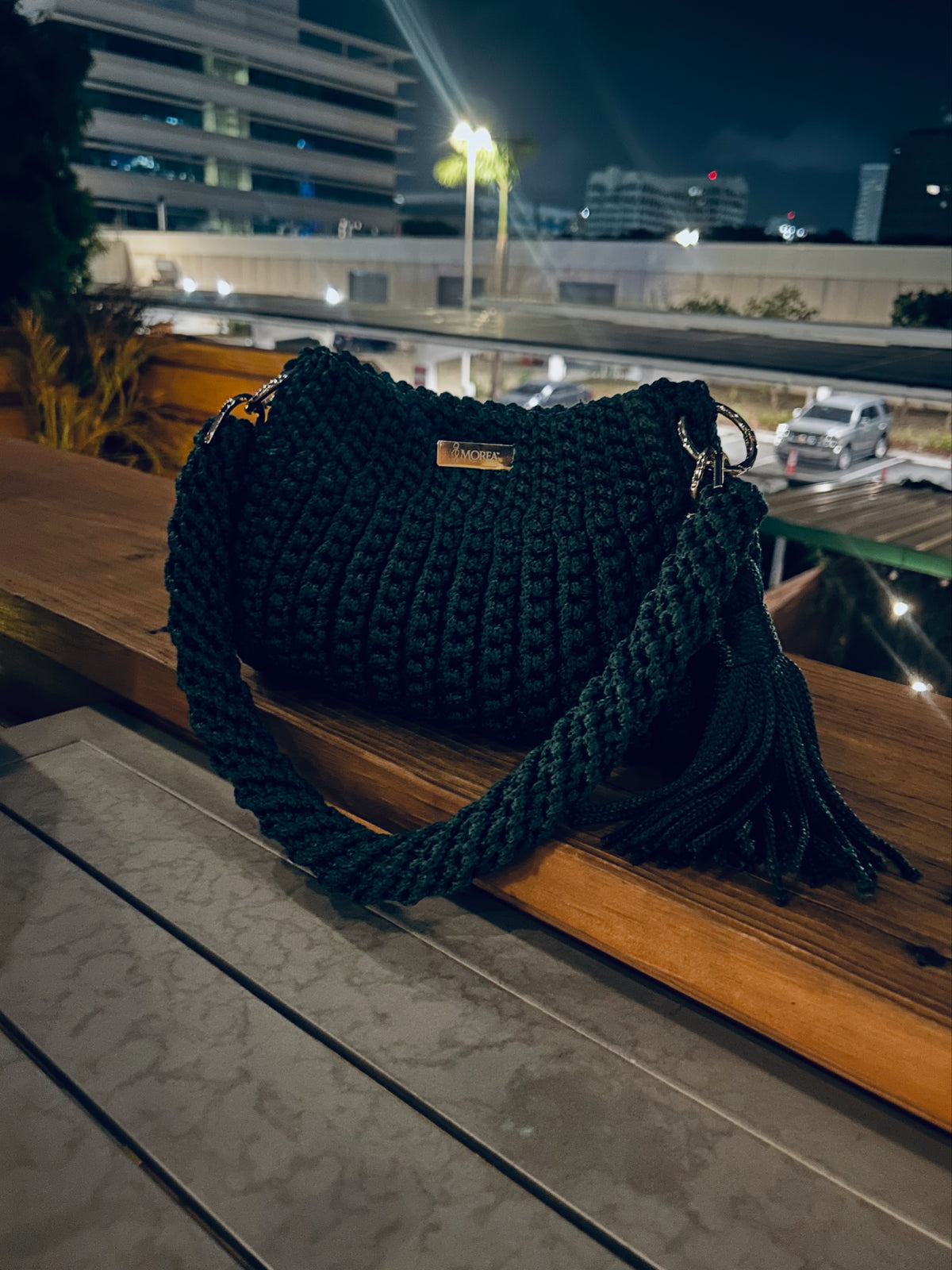 Dark green crochet Jolie shoulder bag with a fringe tassel, photographed outdoors at night with a city skyline in the background.