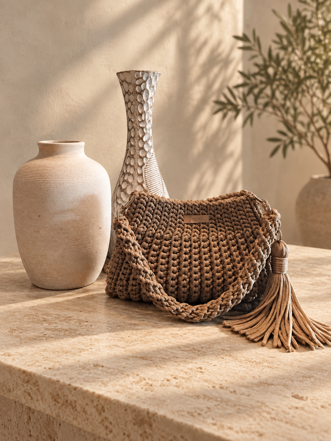 Tan crochet Jolie shoulder bag with a fringe tassel, displayed on a stone table beside ceramic vases.