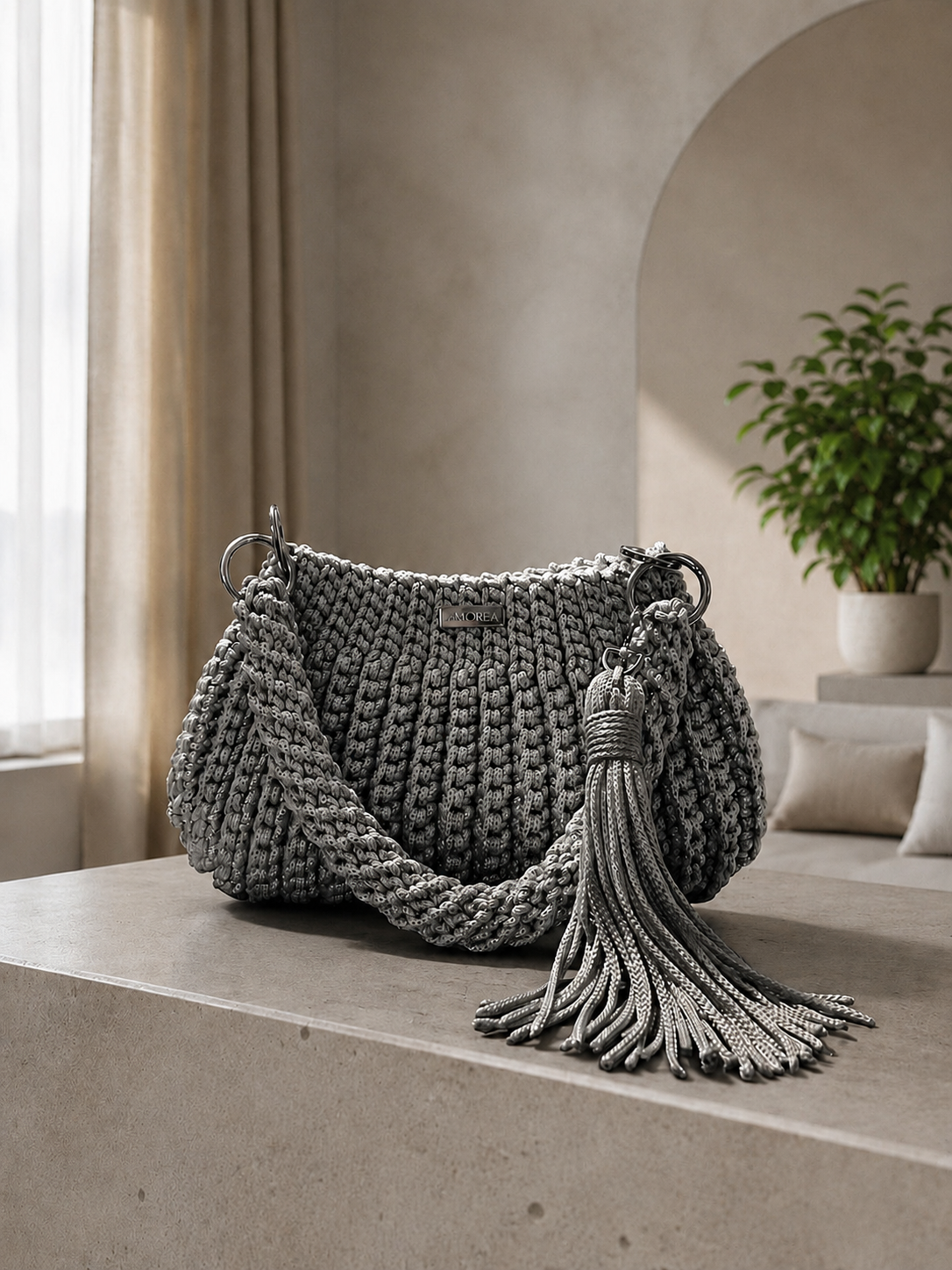 Grey crochet Jolie shoulder bag with a fringe tassel, displayed on a pedestal beside a potted plant and archway.